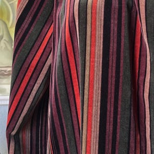 May include: A velvet garment with vertical stripes in shades of red, black, brown, and green. The garment has a high neckline and a button closure. The sleeves are long and the overall style is vintage.