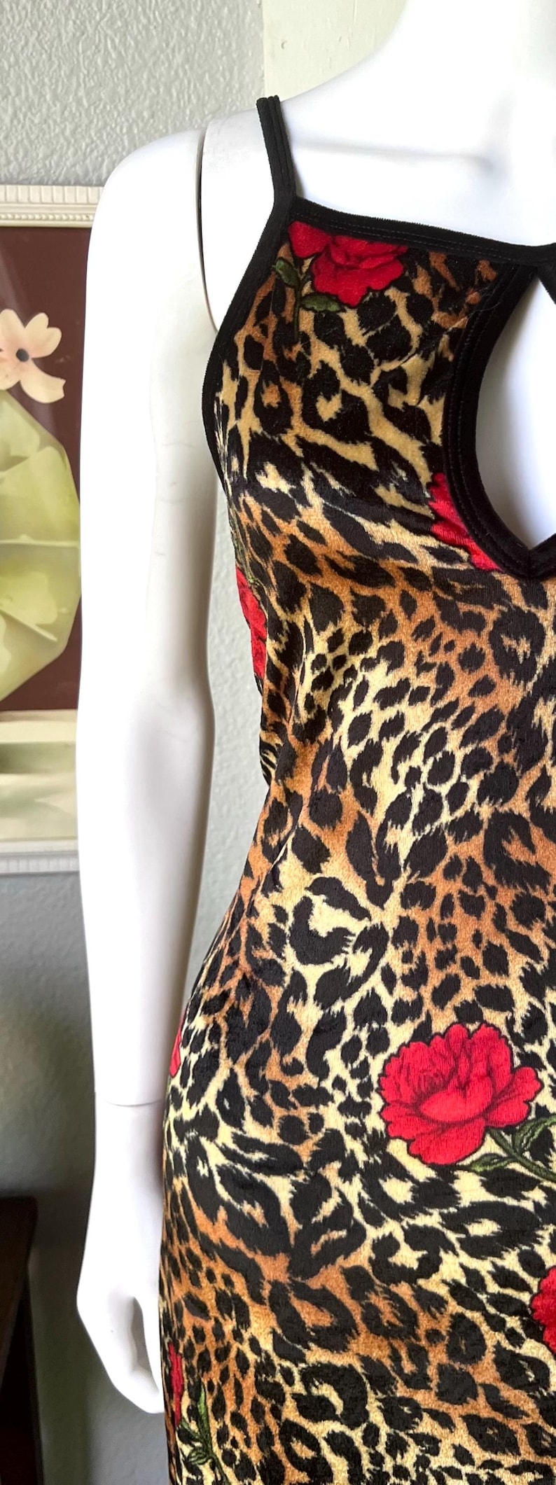 May include: A sleeveless dress with a leopard print pattern in shades of brown and black. The dress features red rose accents and a black trim. The garment is displayed on a mannequin.