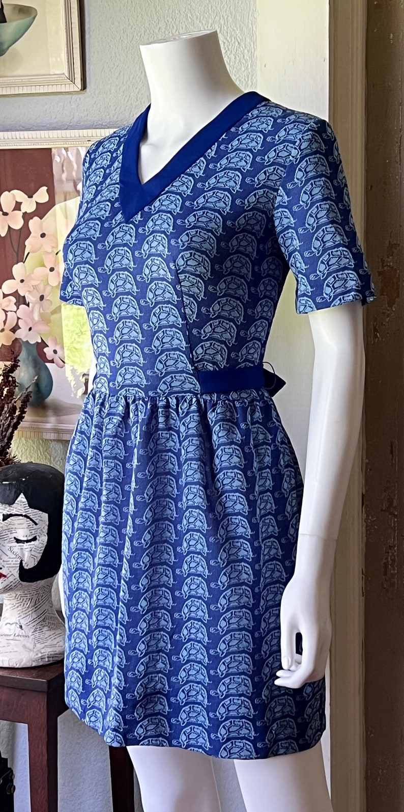 May include: A blue and white short-sleeved dress with a turtle print. The dress has a v-neck and a tie at the waist.