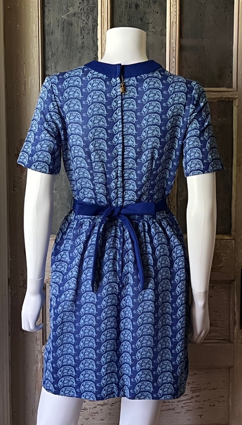 May include: A blue short-sleeved dress with a white turtle print. The dress has a navy blue sash that ties at the back.