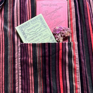May include: A close-up of a striped fabric in shades of red, pink, and black. A pocket holds a green book titled "Shakespeare's Merchant of Venice" and a pink book. A small bouquet of dried flowers rests nearby.