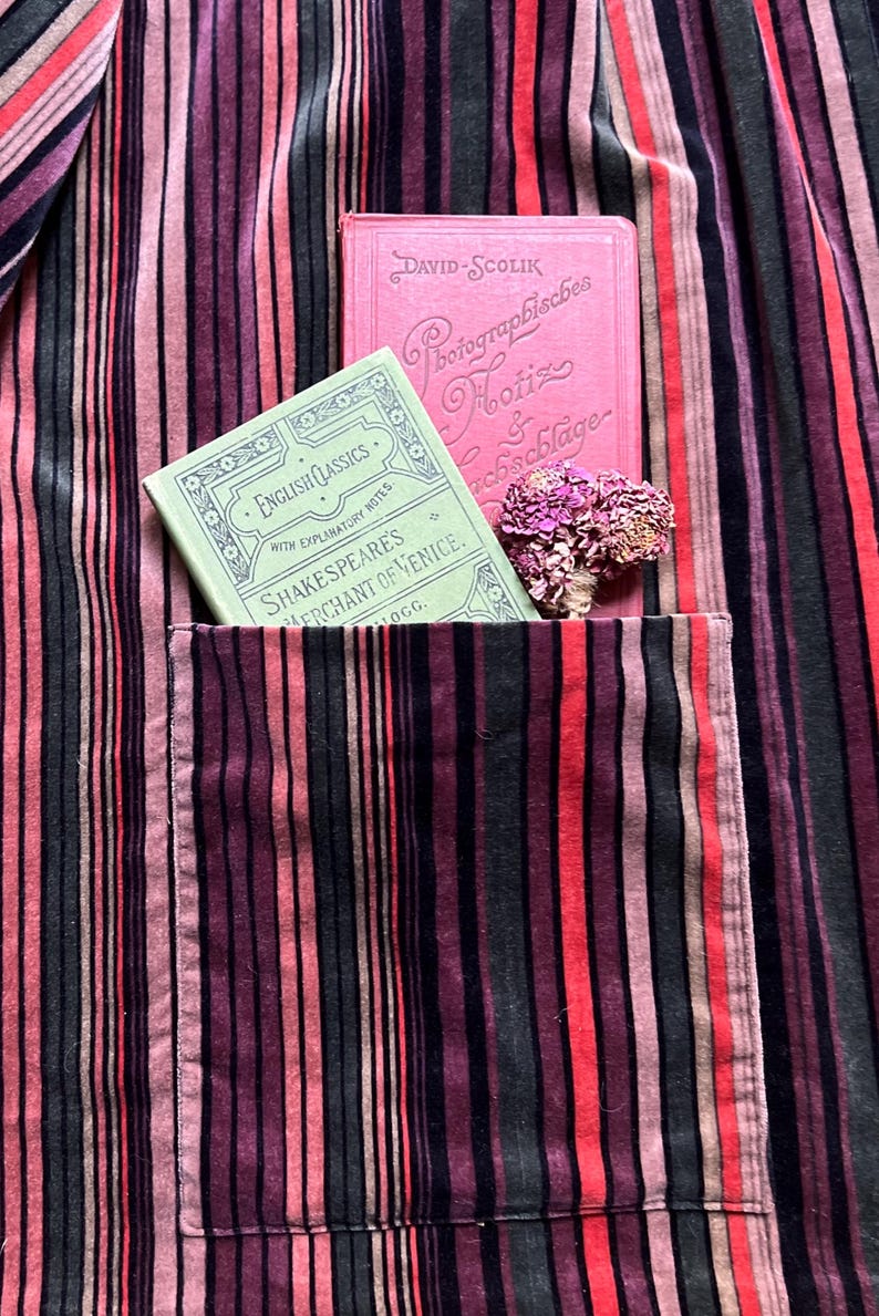 May include: A close-up of a striped fabric in shades of red, pink, and black. A pocket holds a green book titled "Shakespeare's Merchant of Venice" and a pink book. A small bouquet of dried flowers rests nearby.