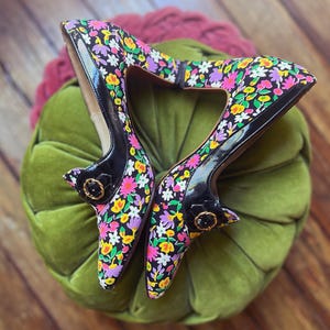 以下が含まれることがあります： 黒いパテントレザーのアクセントが付いた、花柄プリントのハイヒール。ヒールは、黒い背景にピンク、黄色、紫の花のカラフルなパターンが特徴です。靴には小さなバックルのディテールがあります。
