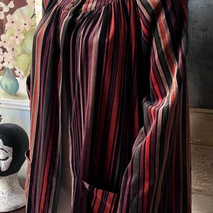 May include: A vintage-style, striped jacket in shades of red, brown, and green. The jacket features vertical stripes, a round neckline, and long sleeves. It has two front pockets and is displayed on a mannequin.