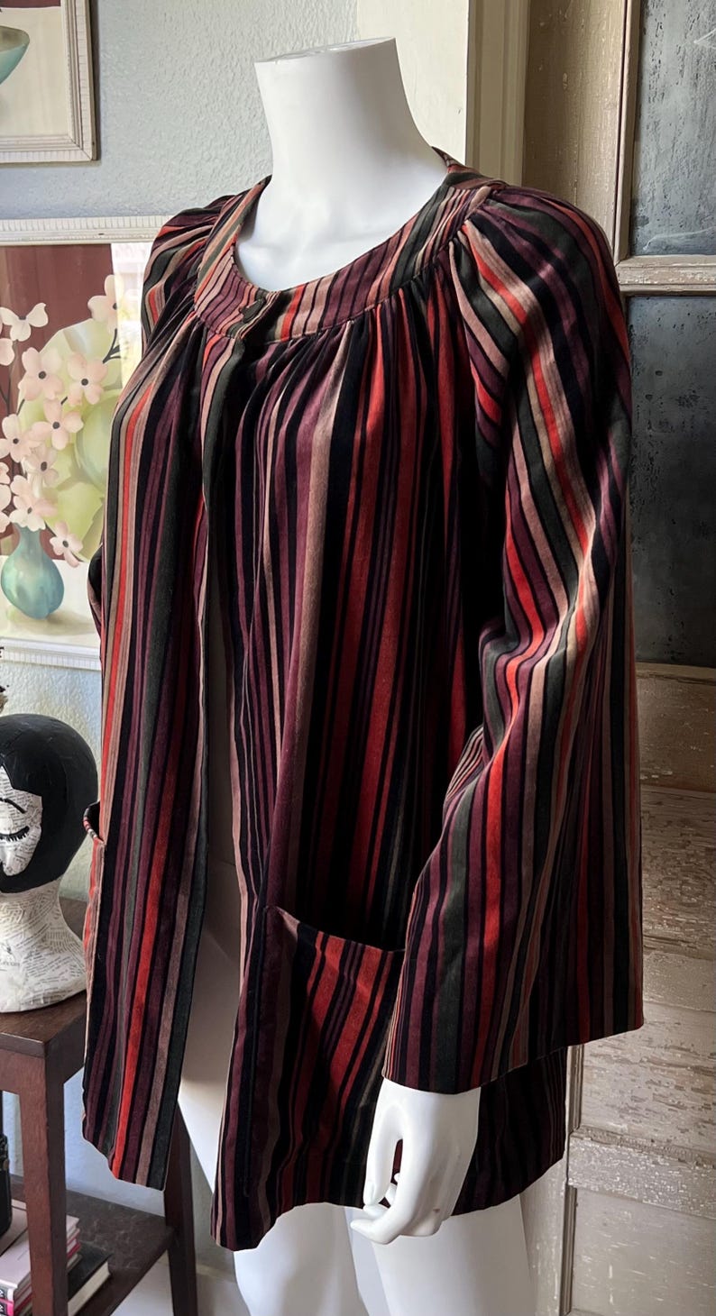 May include: A vintage-style, striped jacket in shades of red, brown, and green. The jacket features vertical stripes, a round neckline, and long sleeves. It has two front pockets and is displayed on a mannequin.