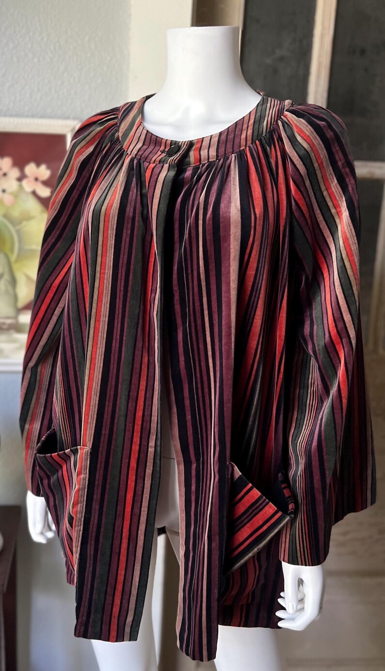 May include: A multi-colored striped jacket with a round neckline and two front pockets. The jacket features vertical stripes in shades of red, green, black, and brown. The garment is displayed on a mannequin.