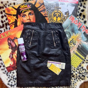 May include: Black leather skirt with silver zippers and stud accents. The skirt is displayed with vintage vinyl records and a can of Aqua Net hairspray. The skirt has a unique design and is a vintage fashion item.