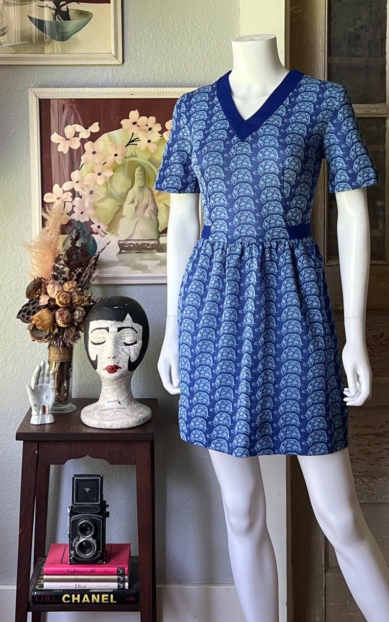 May include: A blue and white short-sleeved dress with a v-neck and a pattern of white turtles on a blue background. The dress has a blue trim around the neckline and waist.