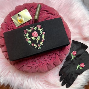 May include: A black rectangular clutch bag featuring a central shield-shaped embroidery of a pink rose surrounded by green leaves and small red flowers, accompanied by a pair of black gloves with a single embroidered pink rose on each wrist.