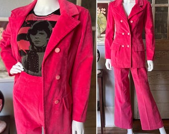 May include: A vibrant pink corduroy suit, consisting of a double-breasted blazer and matching pants, displayed on a mannequin. The blazer has large buttons and a tailored fit. A graphic t-shirt is visible underneath the blazer. The suit is a bold fashion statement.