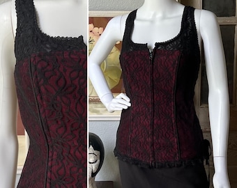 May include: A black and burgundy corset top with a square neckline and a front zipper. The top is made of burgundy fabric overlaid with black lace. The straps and neckline are trimmed with black lace.