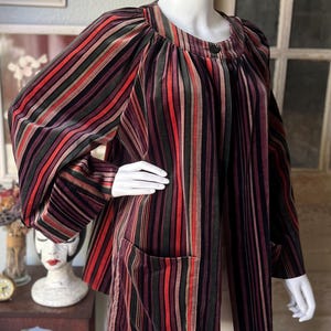 May include: A vintage, striped jacket in shades of burgundy, red, green, and brown. The jacket features a round neckline, long sleeves, and two front pockets. The fabric has a velvet-like texture.