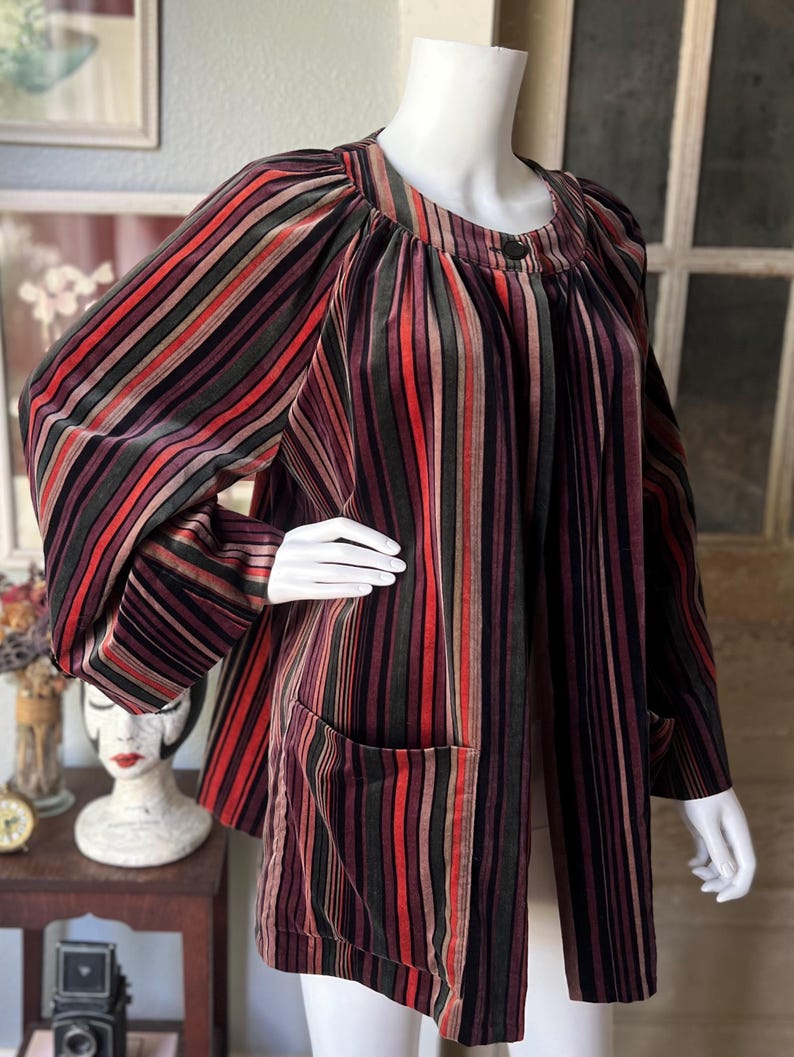 May include: A vintage, striped jacket in shades of burgundy, red, green, and brown. The jacket features a round neckline, long sleeves, and two front pockets. The fabric has a velvet-like texture.