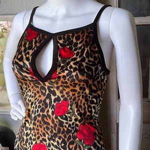 May include: A sleeveless dress featuring a leopard print pattern with red rose accents. The dress has a keyhole neckline and black trim. The garment is displayed on a white mannequin.