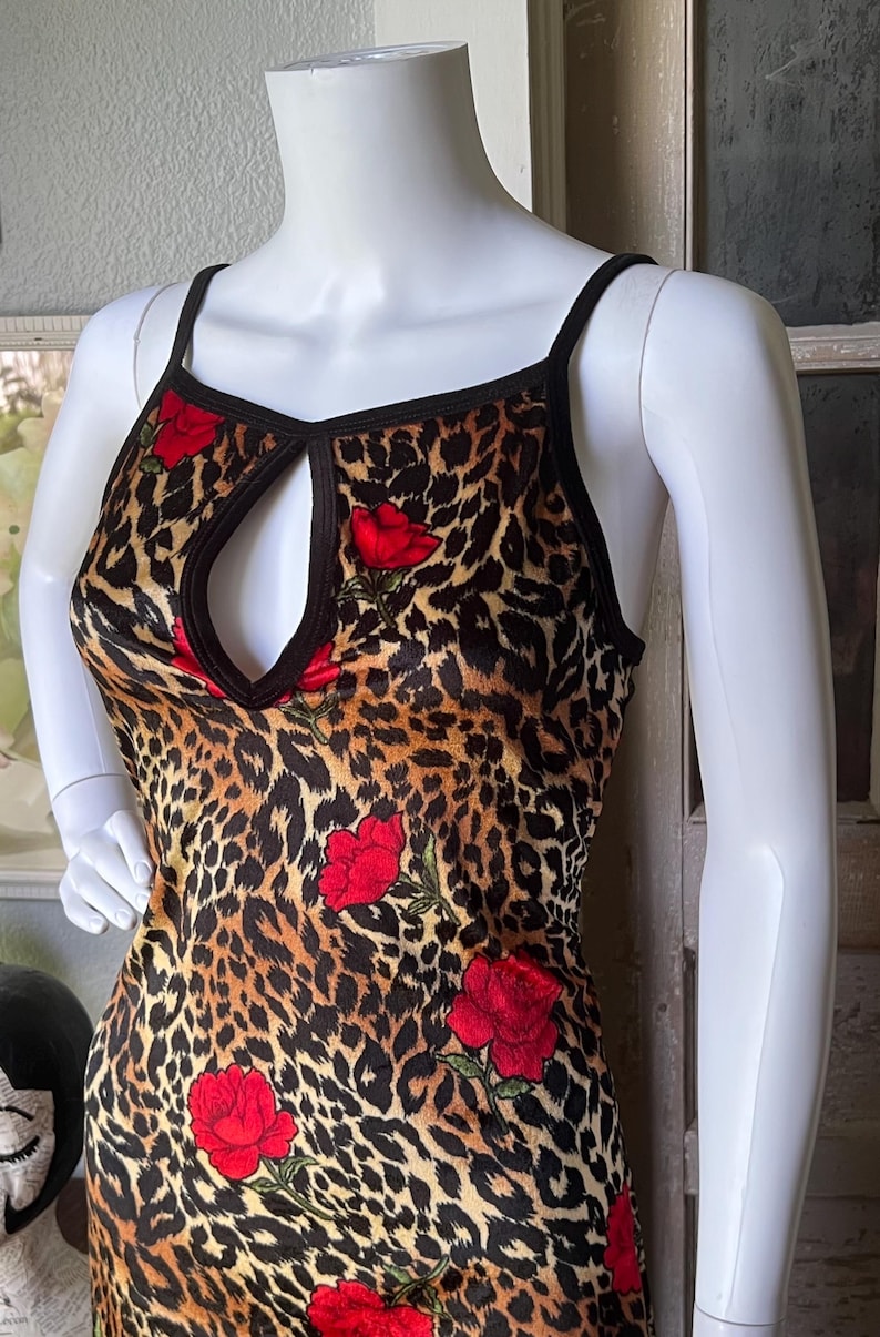 May include: A sleeveless dress featuring a leopard print pattern with red rose accents. The dress has a keyhole neckline and black trim. The garment is displayed on a white mannequin.
