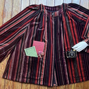 May include: A striped, burgundy velvet jacket with a round collar and long sleeves. The jacket has a pocket with two small books and a small camera. The stripes are in shades of red, black, and brown.