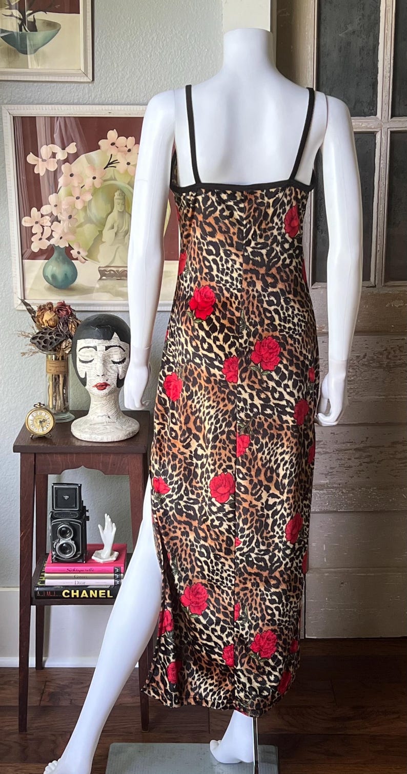 May include: A leopard print dress with red rose accents. The dress has a black trim and spaghetti straps. The dress is displayed on a mannequin. The dress has a slit on the left side.