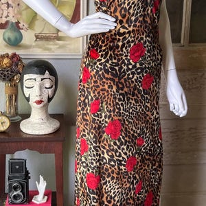 May include: A sleeveless, floor-length dress on a mannequin. The dress features a leopard print pattern with red rose accents. The neckline has a keyhole design and black trim.