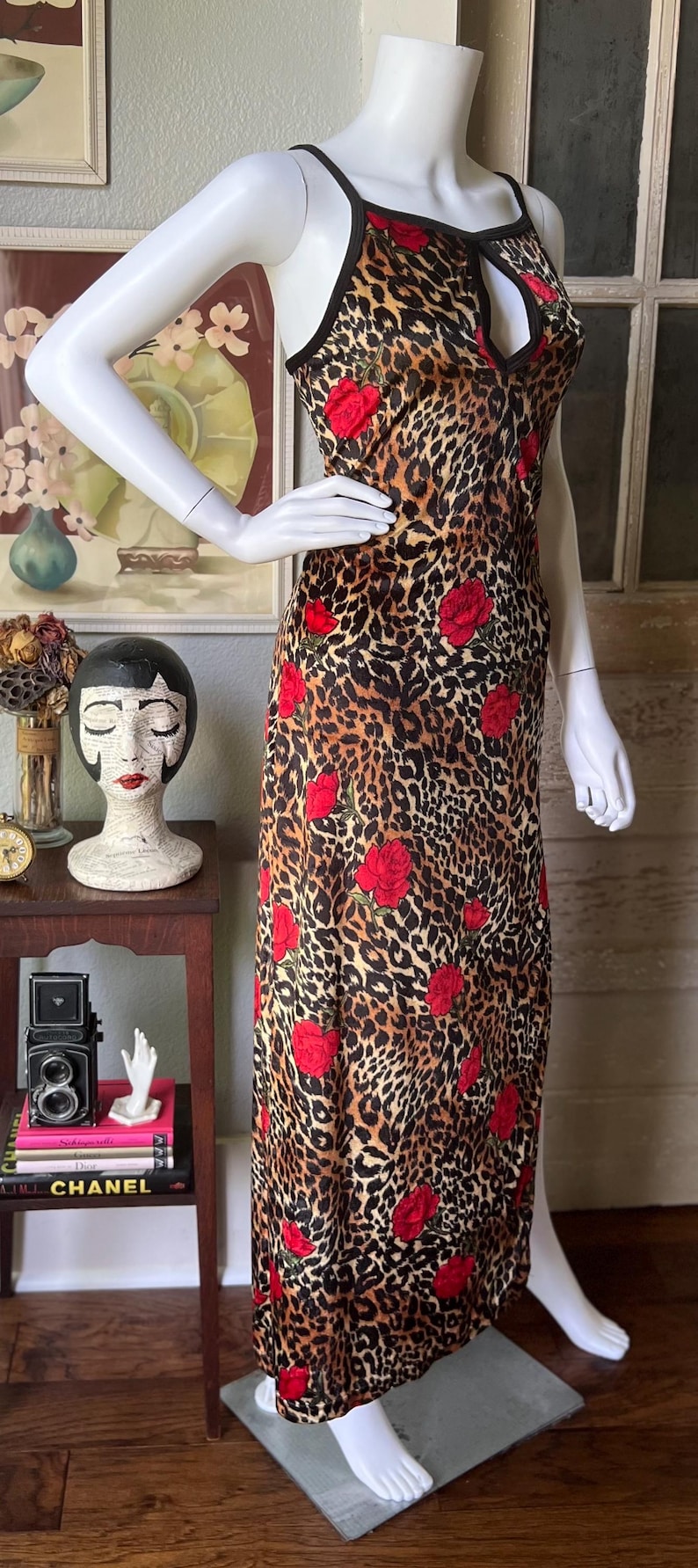 May include: A sleeveless, floor-length dress on a mannequin. The dress features a leopard print pattern with red rose accents. The neckline has a keyhole design and black trim.