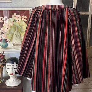 May include: A multi-colored striped cape with a round neckline. The cape features vertical stripes in shades of red, black, green, and brown. The garment is displayed on a mannequin. The cape is made of a soft, flowing fabric.