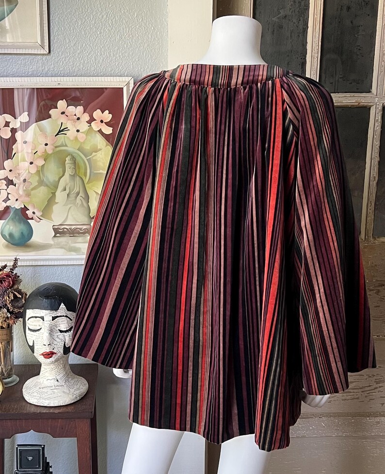 May include: A multi-colored striped cape with a round neckline. The cape features vertical stripes in shades of red, black, green, and brown. The garment is displayed on a mannequin. The cape is made of a soft, flowing fabric.