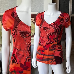 May include: A short-sleeved, V-neck shirt in shades of red, orange, and brown. The shirt features a graphic design of a face with intricate details and gold-colored embellishments. The bottom of the shirt has a patchwork design.