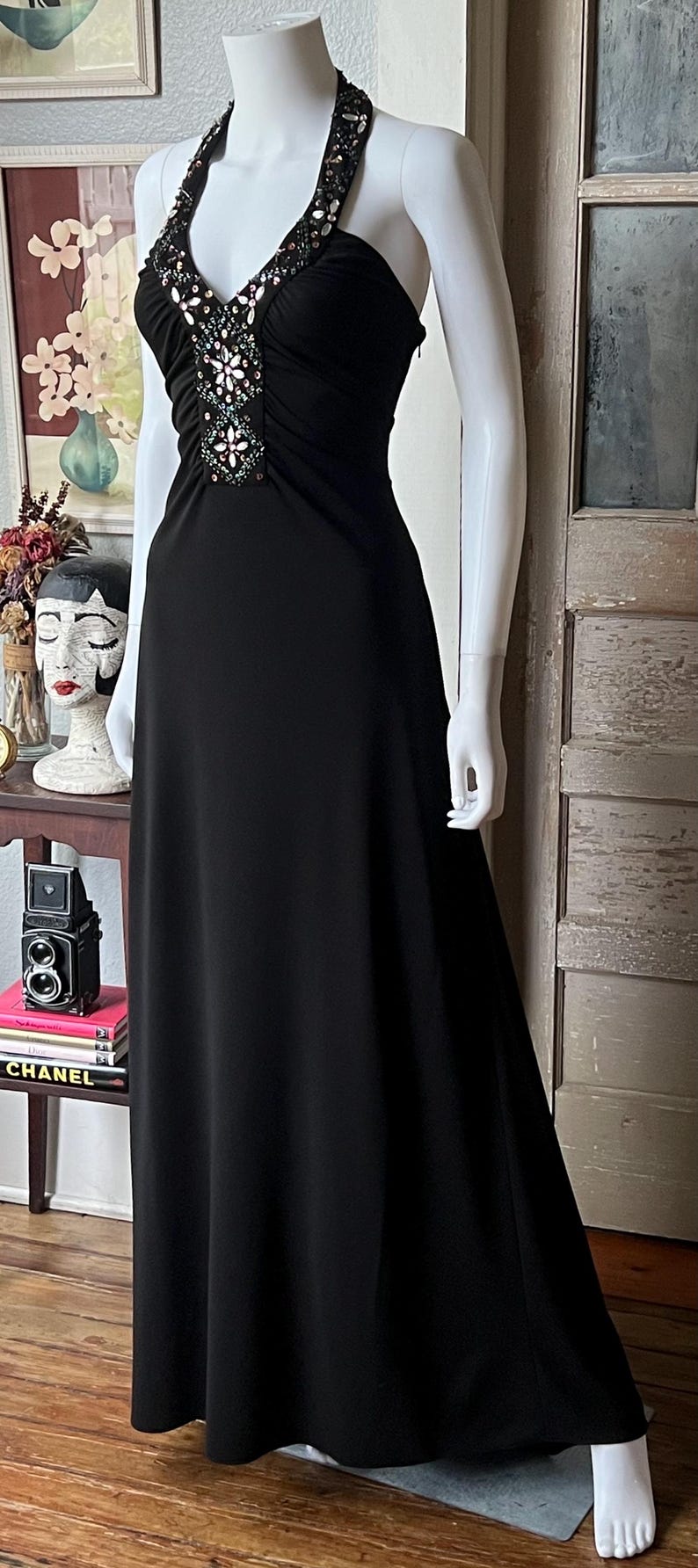 以下が含まれることがあります： ビーズがあしらわれた黒いホルターネックのロングドレス。ドレスは体にフィットしたボディと、ゆったりとしたスカートが特徴です。