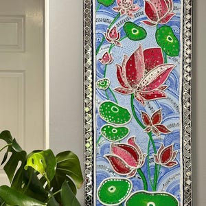 Puede incluir: Una pieza de arte de pared enmarcada que presenta un fondo azul con flores de loto rosas y rojas y hojas verdes. El marco es plateado con un borde de espejo.