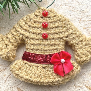 May include: A crocheted sweater ornament with a red glitter belt and a red bow with a pearl center. The sweater is a light brown color with red buttons.