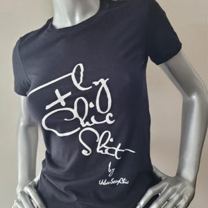 Può includere: Maglietta nera con testo bianco che dice "Fly Chic Shit" di Urban Sexy Chic.