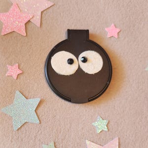 May include: A black, round object with white, glittery eye shapes and black pupils, resembling a cartoon face. Scattered around are various glittery star shapes in pink and green hues, all set against a neutral background.