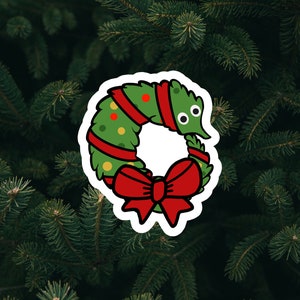 May include: A whimsical Christmas wreath sticker with a cartoon worm design. The wreath is green with red ribbon and a large red bow. The background is a dark green, textured surface of evergreen branches.