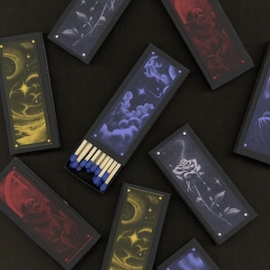 May include: A collection of decorative matchboxes with various designs. Each box features a different illustration, including a rose, clouds, a crescent moon, and a grim reaper. The matches have blue tips and are displayed on a black surface.