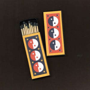 May include: Two matchboxes with a yin-yang design. One box is open, revealing the matchsticks with blue tips. The boxes have a navy blue background with a yellow border and a red and white yin-yang symbol. The design includes small star accents.