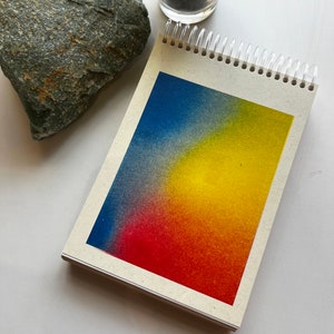 May include: A small, spiral bound notebook with a gradient background. The background is a blend of blue, yellow, and red colors.