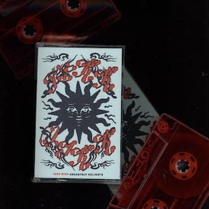 May include: A clear cassette tape with a white label featuring a black sun design and red text. The text reads "TEKK NIKK UNEARTHLY DELIGHTS". Two additional red cassette tapes are visible in the background.
