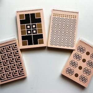 May include: Four spiral-bound notepads with geometric designs. The covers are a light brown color with black and gold patterns. The designs include squares, intersecting lines, and circular shapes. The spiral binding is white.