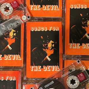 May include: A stack of cassette tapes with orange and black labels. The labels feature the text "Songs for the Devil" and an image of a person with a mask. The tapes are in clear plastic cases.