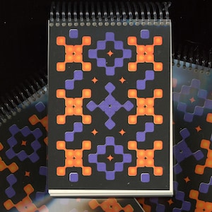 May include: Spiral-bound notepad with a black cover featuring a geometric pattern in orange and purple. The design is repeated on the cover of other notepads in the image. The pages are white.