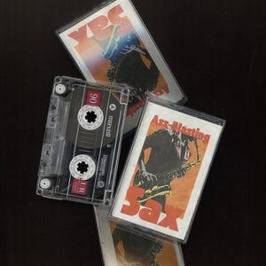 May include: A vintage cassette tape with a clear plastic case and a black label. The label says "Maxell UR 90" and has a red "A" side indicator. The cassette is sitting on top of two other cassette tapes. The tape on the left has a red and yellow label with the text "Yes" and the tape on the right has a red and orange label with the text "Ass-Blasting Sax".