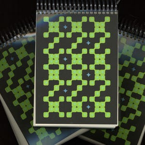 May include: Spiral-bound notepad with a black cover featuring a repeating pattern of neon green squares and blue star shapes. The notepad is open, revealing the patterned design on the pages. Additional notepads are visible in the background.