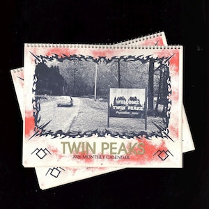 May include: A 2026 monthly calendar featuring a black and white image of a road leading to a "Welcome to Twin Peaks" sign. The calendar has a red border and the words "Twin Peaks" in gold lettering.