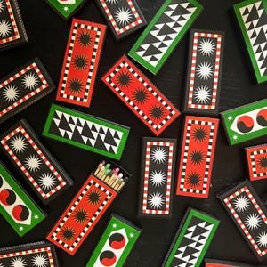 May include: An assortment of decorative matchboxes with geometric designs. The matchboxes feature red, green, and black backgrounds with white starbursts, triangles, and other patterns. One box is open, revealing the matches inside.