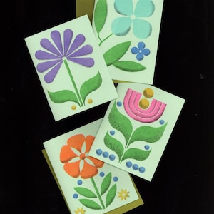 May include: Four greeting cards with floral designs. Each card features a different flower in shades of purple, blue, orange, and pink, with green leaves and stems. The cards are on a light green background with gold-colored envelopes.