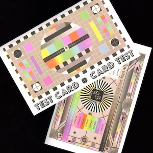 May include: Two white test cards with colorful patterns and text. The cards feature a circular color bar pattern, squares, and text that reads "TEST CARD". The cards are designed for calibration and testing.
