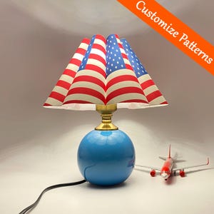 Puede incluir: Lámpara de mesa decorativa con base esférica azul y pantalla con diseño de la bandera estadounidense. La lámpara tiene un cuello color latón y un cable de alimentación negro. La imagen también incluye un pequeño avión de juguete rojo y blanco.