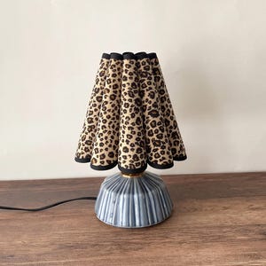 May include: A table lamp featuring a pleated fabric lampshade with a tan and black leopard print pattern and black trim. The base is made of glossy, light blue ceramic with a vertical ribbed texture and a gold-colored metal neck connector.