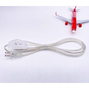 May include: A clear lamp cord with a clear inline switch and a two-prong plug. The cord is looped and secured with a small blue band. A red and white toy airplane is in the background.
