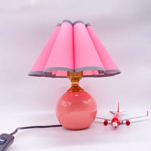 May include: A pink ceramic table lamp with a round base and a pleated lampshade. The lampshade is pink with a gray trim. A red and white toy airplane sits next to the lamp. The lamp has a gold-colored neck.