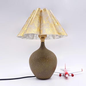 May include: A decorative table lamp with a gold and white patterned lampshade. The lamp has a dark brown, textured base and a brass neck. A small red and white toy airplane is next to the lamp.